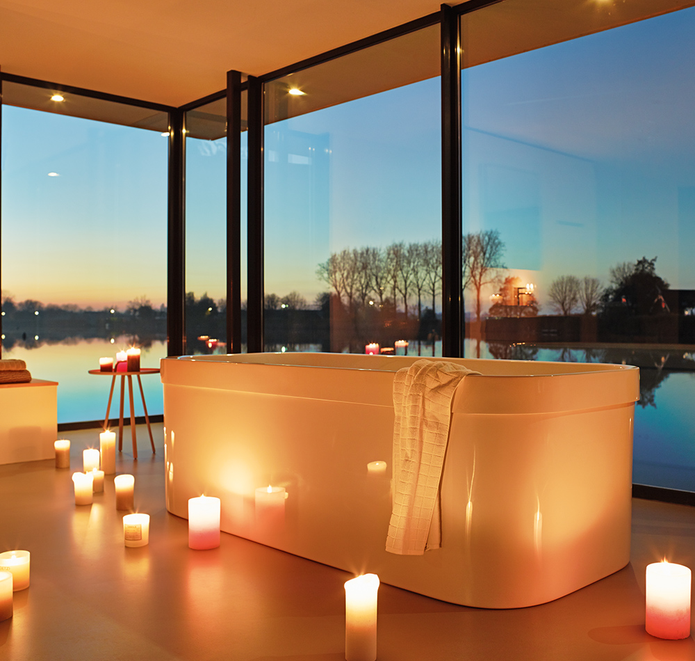 Ein stimmungsvolles Wellness-Ambiente mit einer freistehenden weißen Badewanne im Mittelpunkt.
Sie ist von zahlreichen warmglühenden Kerzen umrahmt, die eine entspannende Atmosphäre schaffen. Durch die bodentiefen Fenster eröffnet sich ein atemberaubender Ausblick auf einen See bei Sonnenuntergang, mit kahlen Bäumen, die sich am Horizont abzeichnen. Die Kombination aus natürlichem Abendlicht und dem warmen Kerzenschein erzeugt eine besonders romantische und luxuriöse Atmosphäre. Ein weißes Handtuch liegt elegant über dem Rand der Badewanne drapiert.
