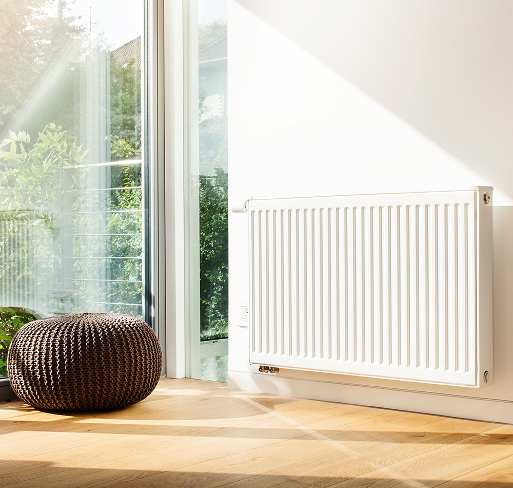 Helles, modern eingerichtetes Zimmer mit großem Fenster, durch das Sonnenlicht hereinfällt. An der weißen Wand befindet sich ein klassischer, weiß lackierter Heizkörper mit vertikalen Rillen.
Auf dem hellen Holzfußboden steht ein runder, brauner Pouf aus geflochtenem Material. Draußen vor dem Fenster sind grüne Pflanzen und ein Balkongeländer zu sehen.