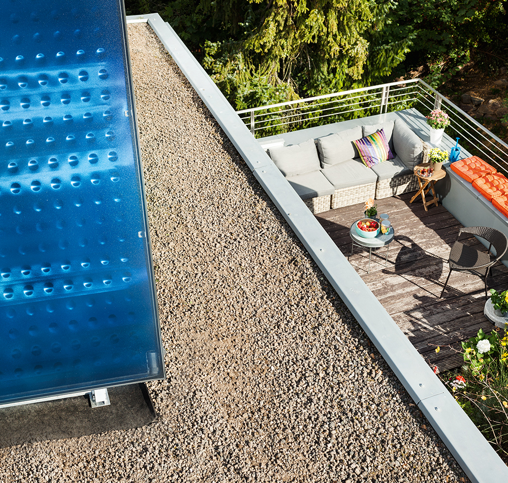 Eine moderne Terrasse mit gemütlicher Lounge-Atmosphäre, aufgenommen aus der Vogelperspektive. Im Vordergrund ist ein blaues Solarpanel mit strukturierter Oberfläche zu sehen, daneben ein Kiesweg.
Die Terrasse selbst besteht aus Holzdielen und ist mit einer grauen Rattan-Sitzgruppe ausgestattet, die helle Polster und ein buntes Dekokissen hat. Ein kleiner Beistelltisch und Dekoelemente wie Pflanzen und Accessoires runden das einladende Ambiente ab. Die Terrasse wird von einem modernen Metallgeländer umrahmt und ist von üppigem Grün umgeben.